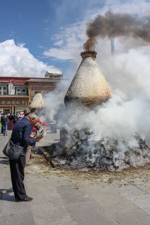 Smoking sangkang incense burner on Barkhor square with a man pilgrim wearing shaggy cloak in a bow with hat offの写真素材