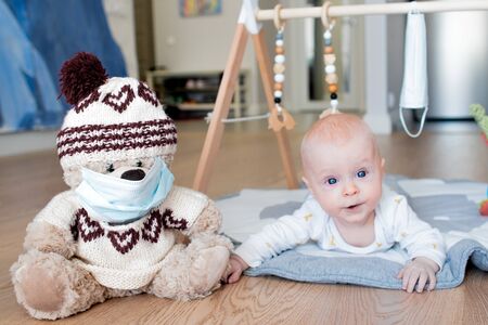 Teddy bear toy wearing a blue face mask and a baby boy lying on the floor and reaching towards his toyの写真素材