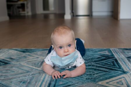 White caucasian baby boy in a face mask lying on the blue geometric carpet in an apartment looking at the camera with mask and head downの写真素材