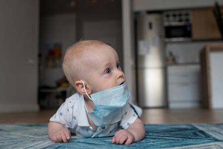 White caucasian baby boy in a face mask lying on the blue geometric carpet in an apartment looking...