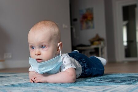 White caucasian baby boy in a face mask lying on the blue geometric carpet in an apartment looking...