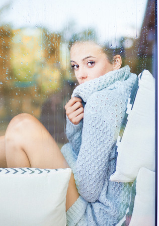 Beautiful young  brunette woman wearing knitted dress sitting home behind a window covered with rain drops. Blurred fall garden reflection on the glass.の写真素材