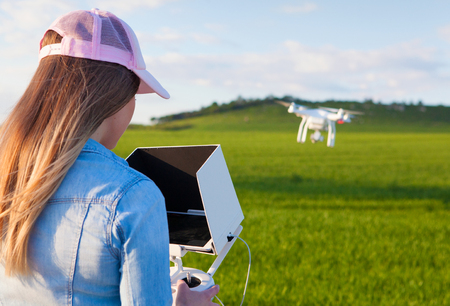 Young woman holding radio controller with tablet and sun shade flying drone uav over a field. Aerial video and photography maker. Hovering aircraft in the background.の写真素材