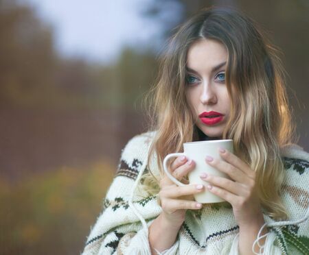 Young beautiful blonde woman with cup of coffee standing by the window. Garden reflection on the glassの写真素材
