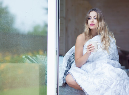 Young beautiful blonde woman with cup of coffee sitting home on the bed covered with duvet by the window. Lazy morning day off conceptの写真素材