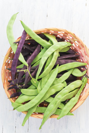 Green and purple beans in a wicker basket on a white wooden backgroundの写真素材