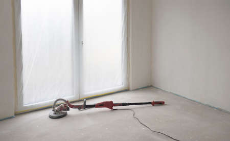 Dirty and dusty red sander for walls and ceilings lies on the floor in empty room against white ready-to-paint walls and floor-to-ceiling French window.Concept of repair of apartment, building house.の写真素材