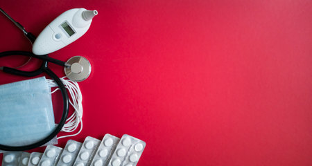 Stethoscope, pack of medical masks, electronic ear thermometer, row of antibiotic pills in blister packs on hue red background on left.Copy space.Prevention and diagnosis of coronavirus.Pandemic concept.の写真素材