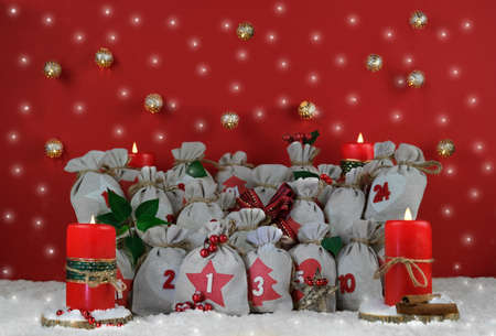 Traditional Advent calendar sacks of gray canvas tied with twine,4 candles in corners,Christmas decor on red with many lights.Red berries,wooden star,Poinsettia leaves,snow.Celebration gifts conceptの写真素材
