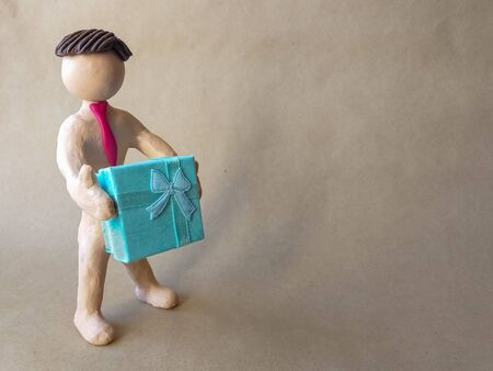 Plasticine figure of a man in a tie who holds a gift in the form of a light blue box with a ribbon. Beige kraft paper background. Illustration for a gift February 14th, birthday. place for text.の写真素材