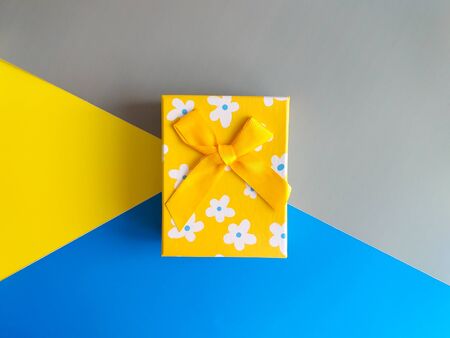 Yellow gift box tied with a gold ribbon. Top view on a contrasting background of bright blue, yellow and gray paper lying diagonally. Place for text, postcards. Abstract background for design.の写真素材