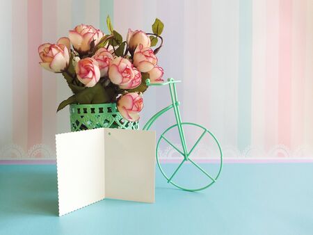 Against the background of striped wallpaper in pink pastel colors, there is a decorative bicycle with flowers. and an open card for congratulations. Space for text, congratulations. Exquisite background to create a summer mood.の写真素材