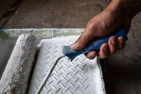 Hand of a house painter with a paint roller. Paint roller with white paint in hand, lowers into a tray with paint, on a concrete floor backgroundの写真素材