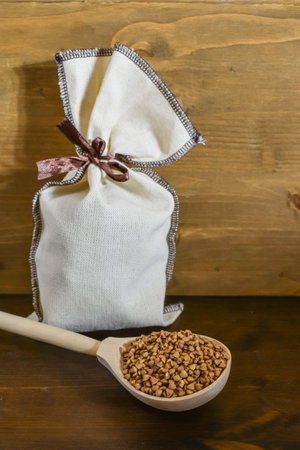 Buckwheat in a wooden spoon, next to a canvas sac with grain on a brown wooden background. Healthy food concept, vegetarian food. Copy space. Selective focusの写真素材