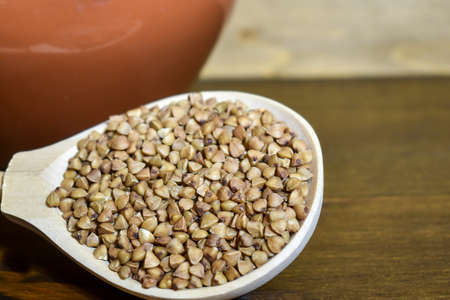 Buckwheat in a wooden spoon, next to a clay pot on a brown wooden background. The concept of healthy dietary food, vegetarianism, traditional Russian cuisine. Selective focus, place for textの写真素材