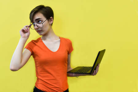 A young woman with short hair, wearing round glasses, holds a laptop in her hands and looks to the side. Student working at the computer. Place for your text. Selective focus.の写真素材