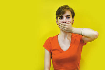 A young woman with short hair covered her mouth with her hands. Isolated on a yellow background. Keep it secret. space for text. Concept: silence, no talking, I won't say anything. Selective focusの写真素材