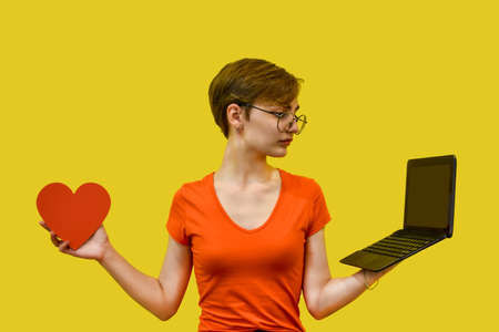 A young woman with glasses makes a choice, holding a red heart in one hand and a laptop in the other. Isolated on a yellow background. Concept: love, technology, long distance relationships, learningの写真素材