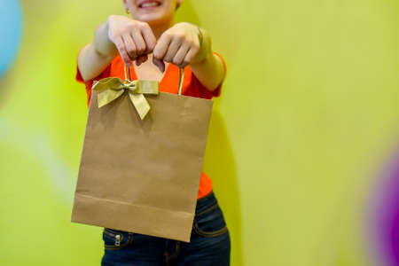 Young woman smiling and holding out a gift bag made of kraft paper against the background of a yellow wall. Concept: birthday gift, purchase, delivery, surprise. Selective focus. Space for text, logo.の写真素材