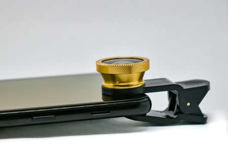 Black smartphone with lens on clip isolated on white. An external wide-angle lens and clip are supplied with the mobile phone. A modern smartphone with an extra lens. Close-upの写真素材