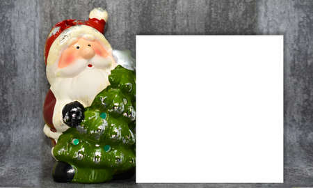 A funny statuette of Santa Claus with a Christmas tree holds a postcard with a space for text. Santa Claus with a white field for text on a gray background. Christmas card.の写真素材