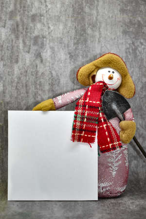 Funny snowman with shovel, on gray background, holds poster with space for text. New Year card with snowman. Smiling snowman holding blank white sheet for text.の写真素材