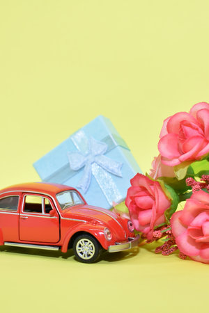 Miniature red car, flowers and gift box. Red retro car delivers bouquet and gift box on yellow background. Valentine, postcard for March 8, Mother's Day, birthday.の写真素材
