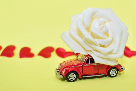 Red car with rose flower on yellow background with red hearts. Red retro car delivers flowers. Valentine, postcard March 8, Mother's Day, birthday. Flower delivery.の写真素材
