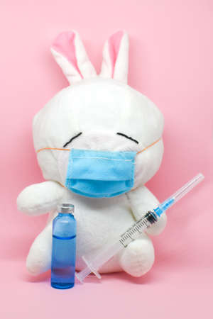 White toy bunny in medical mask with an ampoule and syringe on pink background. copy space, space for text.の写真素材
