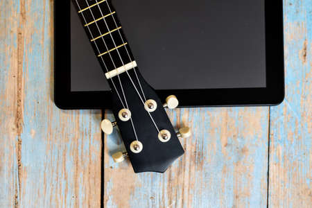 Tablet and ukulele on wooden table with space for text. Online learning to play Hawaiian guitar. ukulele fretboard on textured wooden background next to tablet screenの写真素材