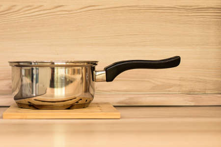 stewpan on wooden table top. A metal stewpan made of stainless steel against background of beige wooden wall and light table top. Kitchen appliances, food preparation. Space for text.の写真素材