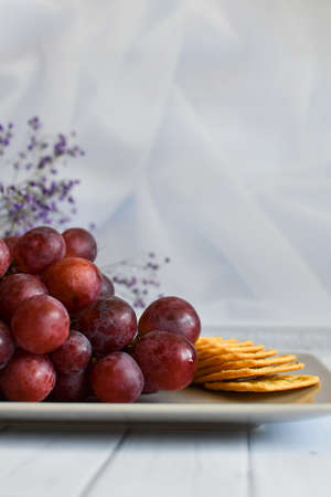 bunch of grapes and cracker on plate. Still life with snack for wine. Red grapes on beige platter on light background. sweet grapes on plate with cheese crackers on white background, space for textの写真素材