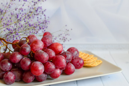 bunch of grapes and cracker on plate. Still life with snack for wine. Red grapes on beige platter on light background. sweet grapes on plate with cheese crackers on white background, space for textの写真素材