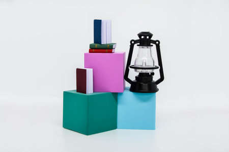 Abstract composition with miniature books and an antique black kerosene lamp standing on a podium of blue and pink cubes. Isolated over white background. Concept: reading, poetry, writing, teaching.の写真素材