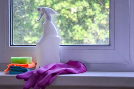 Spray bottle with window cleaner, rag and cleaning sponge lie on windowsill against backdrop of dirty plastic window. Cleaning service concept, cleaning accessories, window washing, tidying upの写真素材