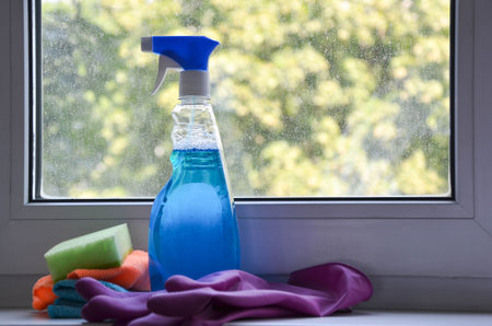 Spray bottle with window cleaner, rag and cleaning sponge lie on windowsill against backdrop of...