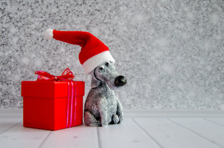 cute funny toy dog in Santa Claus hat sits near red gift box against shiny gray wall background. Postcard for new year, christmas, calendar, poster. space for text.の写真素材