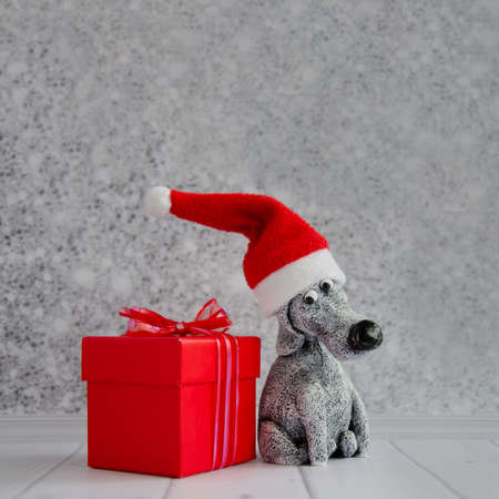 cute funny toy dog in Santa Claus hat sits near red gift box against shiny gray wall background. Postcard for new year, christmas, calendar, poster. space for text.の写真素材