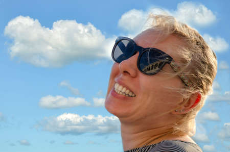 Close-up portrait of a happy laughing woman with sunglasses on the background of a bright blue summer sky with white clouds.の写真素材