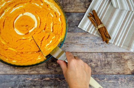 female hand takes a cut piece of pumpkin-curd casserole with a spatula. Festive diet pie. Pumpkin cheesecake on a textured wooden tabletop background. Top view, selective focusの写真素材
