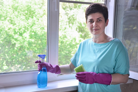 woman with short black hair in rubber gloves washes window in her apartment with rag while holding...