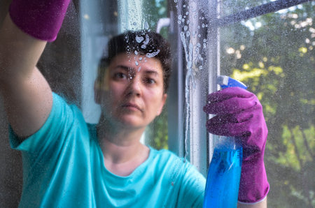 woman with short black hair in rubber gloves washes window in her apartment with rag while holding window cleaner. concept of cleaning services, window washing, housework.の写真素材