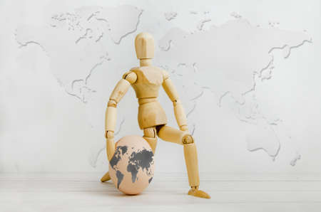Wooden figurine of man holding an egg in form of planet earth against backdrop of world map. concept of environmental protection, ecology, travel, knowledge of the world. Earth day, study of geographyの写真素材