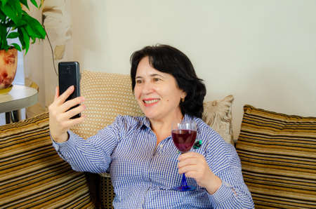 Attractive mature woman holding a glass of wine and talking on the phone while sitting on a sofa in a cozy living room at home. happy elderly woman calling friends via video callの写真素材