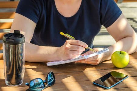 woman makes notes in notebook during lunch, sitting on veranda on sunny day. Apple and Coffee in tumbler on tableの写真素材