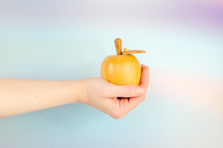 Wooden apple in an outstretched female hand on a blue background. Close-up, woman's hand holding an apple. Concept: temptation, knowledge, offerの写真素材