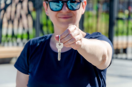 happy woman demonstrates the keys in her outstretched hand. A smiling brunette with short hair joyfully holds the keys to a new apartment in her hands against the backdrop of new buildingsの写真素材
