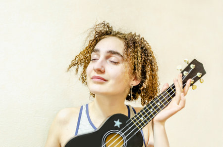 Portrait of a cute funny girl with tousled curly hair playing the ukulele. Young smiling woman holding ukulele in her hands, closeupの写真素材