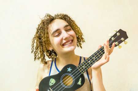 Portrait of a cute funny girl with tousled curly hair playing the ukulele. Young smiling woman holding ukulele in her hands, closeupの写真素材