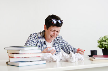 A woman with short black hair at her desk with books, textbooks and crumpled sheets of paper. Creative crisis, sadness, despair from lack of inspiration, problem solving, learning difficulties.の写真素材